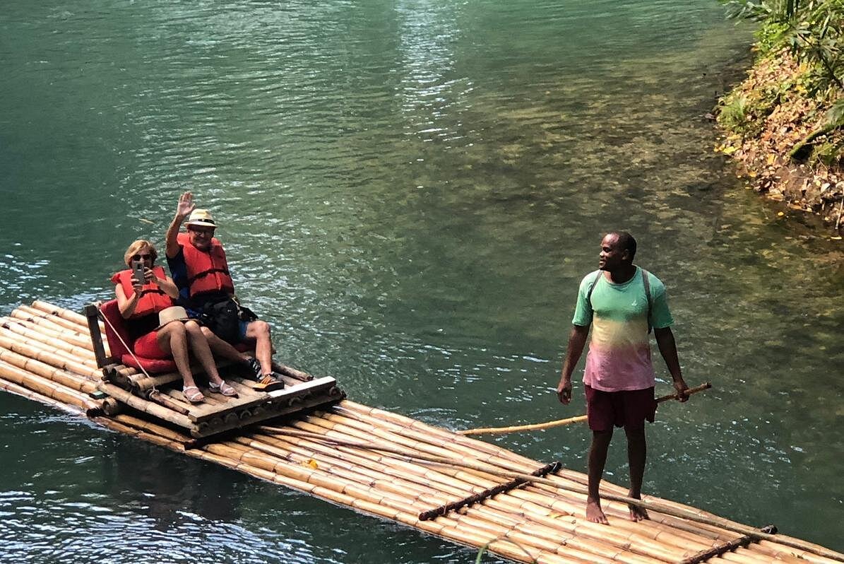 two people sitting on a bamboo raft with life jackets on waving at the person taking the picture and a person guiding the raft in the front